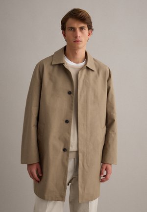 Giovane uomo che indossa un cappotto beige monopetto, un maglione chiaro e pantaloni bianchi, in piedi davanti a uno sfondo grigio chiaro uniforme.