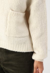 Pull crème tricoté avec une finition texturée, présentant une poche avant et des poignets côtelés, associé à un pantalon en velours côtelé vert foncé.