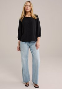 Blusa nera con motivo a pois testurizzati, scollatura rotonda e chiusura con bottoni, abbinata a jeans a gamba larga in denim chiaro e sandali neri.