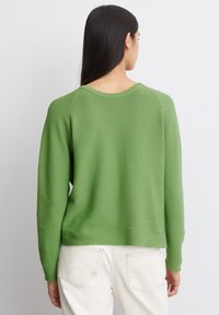 Pull en maille côtelée vert, à col rond, de coupe décontractée et à manches longues, associé à un pantalon blanc. Texture simple sans motifs apparents.