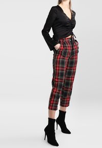 Femme portant un haut noir à manches longues froncées, un pantalon court à carreaux rouge et noir, et des bottines noires à talons hauts, les mains dans les poches.