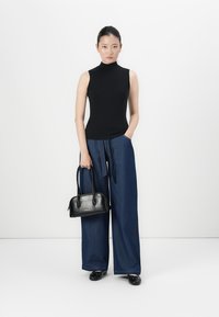 Top preto sem mangas com gola alta, calças largas de ganga azul e uma pequena bolsa preta com duas alças. Modelo de pé num fundo neutro.