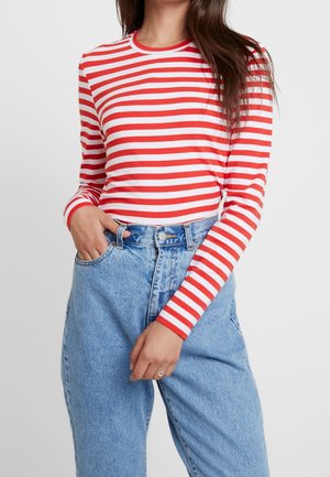 Femme portant un t-shirt à manches longues rayé rouge et blanc rentré dans un jean taille haute bleu clair, avec une main dans la poche.