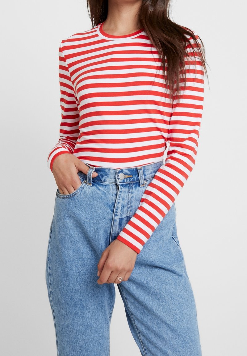 Femme portant un t-shirt à manches longues rayé rouge et blanc rentré dans un jean taille haute bleu clair, avec une main dans la poche.
