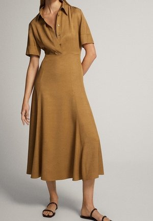 Vrouw in een tan kleurige, kort mouw, knoopsluiting midi-jurk met een col en zwarte sandalen, staande tegen een effen achtergrond.