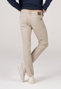 Helle beigefarbene Slim-Fit-Hose aus Baumwolle mit mittelhohem Bund, fünf Taschen und einem braunen Etikett auf der Rückseite. Kombiniert mit weißen Sneakers.