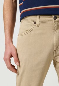 Beżowe dżinsy z pięcioma kieszeniami i widocznym szwem. Materiał wydaje się miękki z delikatną teksturą. Obecny jest brązowy guzik.