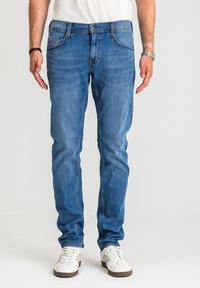 Mężczyzna ubrany w dopasowane niebieskie dżinsy, białe trampki, białą koszulę, czarną opaskę na lewym nadgarstku oraz srebrny zegarek na prawym nadgarstku.