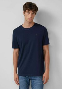 Junger Mann mit lockigem braunem Haar, der ein schlichtes dunkelblaues T-Shirt mit kurzen Ärmeln und helle blaue Jeans trägt, steht vor einer hellgrauen Wand.