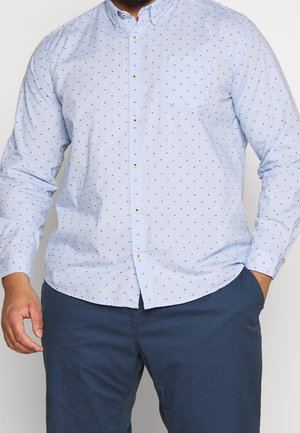 Man wearing light blue button-up shirt with small dark dots and dark blue pants, one hand in pocket and one relaxed by side.
