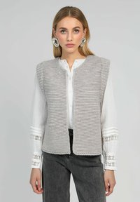 Gilet tricoté gris clair avec un devant ouvert, motif texturé et larges emmanchures, porté sur une chemise blanche à boutons avec des accents en dentelle.