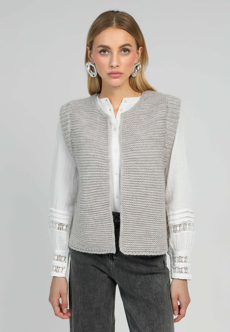 Gilet tricoté gris clair avec un devant ouvert, motif texturé et larges emmanchures, porté sur une chemise blanche à boutons avec des accents en dentelle.