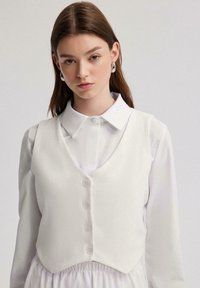 Gilet en lin blanc avec un design à col en V, doté d'une fermeture à boutons sur le devant et d'un ourlet arrondi, superposé à une chemise blanche à col.