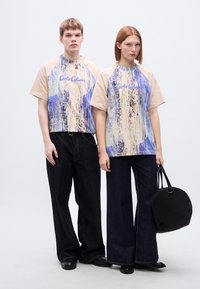 Κοντομάνικα μπλουζάκια με μπλε και μπεζ αφηρημένο σχέδιο, συνδυασμένα με σκούρο τζιν παντελόνι. Ένα άτομο κρατάει μια μαύρη στρογγυλή τσάντα.