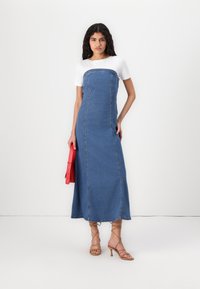 Denim maksi obleka s prilegajočim bodicem in pravim krilom; ima bele kratke rokave, vidno šivanje in rdečo torbico.