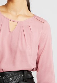 Blusa rosa con scollatura a goccia, pieghe sulla parte anteriore e dettagli in pizzo sulle spalle, abbinata a una cintura nera. Texture liscia.