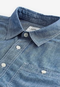 Chemise en denim bleu clair avec col boutonné, boutons blancs, poche avant et détails de couture subtils. Étiquette visible à l'intérieur du col.