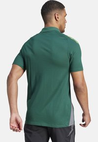 Polo verde con maniche corte, presenta dettagli riflettenti in giallo e un tessuto testurizzato. È realizzato con una vestibilità comoda.