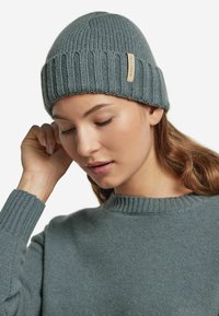 Stickad mössa i mjukt blågrönt ull, med ribbat mönster, vikbar kant och en liten varumärkesetikett på sidan.