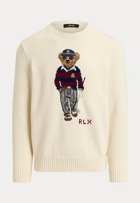 Pull couleur crème avec un ours tricoté portant un maillot de rugby rayé, une casquette, des lunettes de soleil, un pantalon et des chaussures, debout à côté du texte RLX.