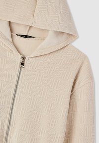 Felpa con zip color beige caratterizzata da un motivo testurizzato, cappuccio e zip argentata. Il materiale sembra morbido con un design tessuto su tutta la superficie.