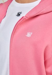 Roze ritsjack met een zachte textuur en een wit logo op de borst. Draagt over een wit t-shirt met een klein zwart logo.
