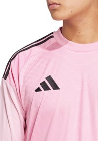 Maglia sportiva rosa chiaro con accenti neri. Presenta un neckline rotondo, tre strisce nere sulle spalle e motivi testurizzati. Logo Adidas in evidenza.