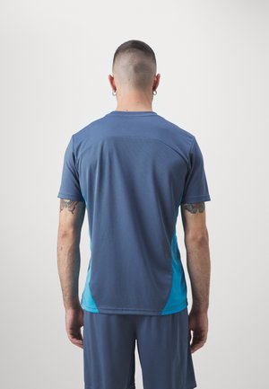 Hombre con cabello corto y pendientes, vistiendo una camiseta deportiva de manga corta azul y turquesa y pantalones cortos a juego, visto de espaldas.