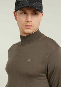Maglione a collo alto marrone a coste con un design aderente e un delicato dettaglio di branding sul petto. Accompagnato da un berretto nero con motivo.