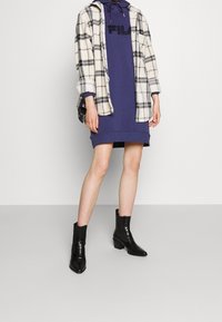 En flanellskjorta i vitt och svart rutmönster, lager på lager över en marinblå klänning. Svarta, texturerade ankelskor med klack fulländar outfiten.