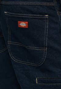 Σκούρο μπλε τζιν παντελόνι με διπλή ραφή στην πίσω τσέπη και μικρή κόκκινη ετικέτα λογότυπου Dickies πάνω από την τσέπη.