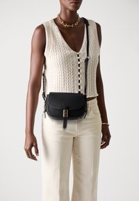 Borsa a tracolla in pelle perforata nera con dettagli in metallo dorato, dotata di chiusura a pattina e tracolla regolabile. Indossata su un abito crema.