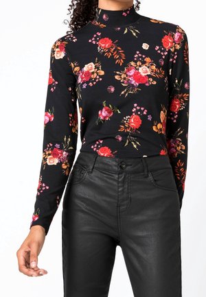 Femme portant un top noir à col roulé à manches longues à motif floral et un pantalon en cuir noir taille haute, debout devant un fond blanc.