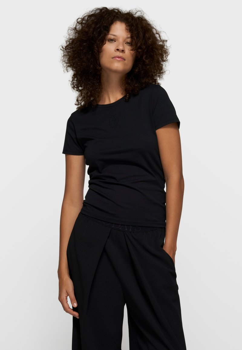 Mujer con cabello rizado, lleva una camiseta negra ajustada de manga corta y pantalones negros sueltos, de pie con una mano en el bolsillo sobre un fondo blanco.