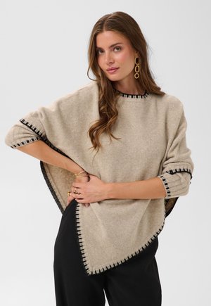 Femme portant un pull beige style poncho avec bordure noire, grandes boucles d'oreilles dorées en forme d'anneau et pantalon noir, posant avec la main sur la hanche devant un fond blanc.