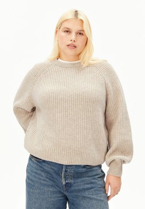 Jeune personne aux longs cheveux blonds portant un pull en tricot beige et un jean bleu, debout devant un fond clair uni.