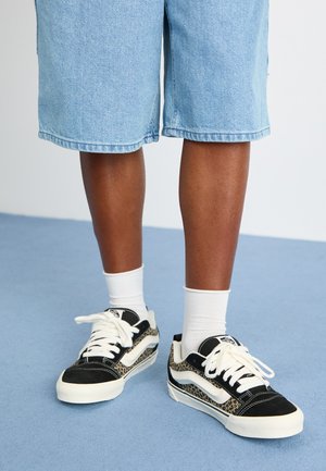 Black and beige sneakers featuring a leopard print design, white laces, and a contrasting white sole, paired with light blue denim shorts.