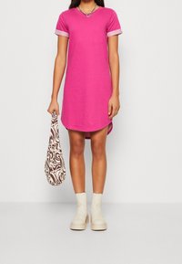 Różowa bawełniana sukienka w stylu t-shirt z krótkimi rękawami, podwiniętymi w jaśniejszym odcieniu. Posiada zaokrąglony dół i jest zestawiona z beżowymi botkami do kostki oraz wzorzystą torbą.