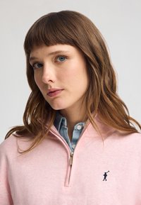 Rosa halvdragspullover i mjukt tyg, med en liten mörk logotyp av en golfspelare på bröstet. Under den finns en ljusblå skjorta med krage.