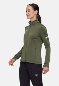 Giacca in pile verde oliva con colletto alto, tasche con zip e una patch con logo sulla manica. Presenta una vestibilità profilata e una texture liscia.
