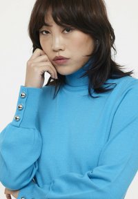 Pull encolure bateau bleu clair en tissu côtelé, avec des détails de boutons sur les manches et une silhouette ajustée.