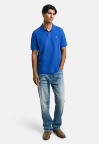 Niebieska koszulka polo z kołnierzykiem i krótkimi rękawami, połączona z jasnoniebieskimi dżinsami i brązowym obuwiem. Koszulka ma małe logo na piersi.