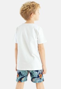 Valkoinen puuvillapaita ja tummansiniset shortsit, joissa on trooppisia lehden ja kukan kuvioita. Shortseissa on mukava istuvuus ja ne on valmistettu kevyestä materiaalista.