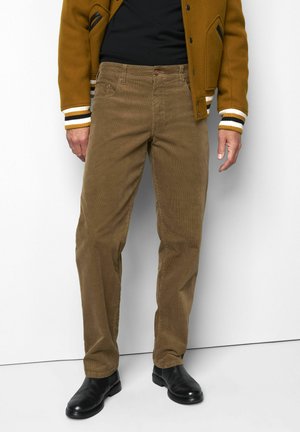 Man draagt een bruine corduroy broek, zwarte schoenen, zwart overhemd en een bruine jas met gestreepte manchetten, staand tegen een witte muur.