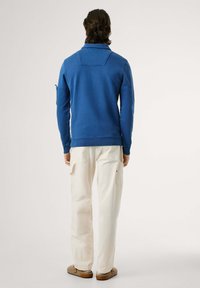 Mėlynas medvilninis džemperis su apykakle ir šoniniu kišeniu, derinamas su kremu cargo kelnėmis su kišenėmis. Matomos rudos neslystančios batus.