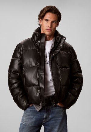 Hombre con cabello castaño que lleva una chaqueta acolchada negra, camiseta blanca y vaqueros azules, de pie con las manos en los bolsillos frente a un fondo liso.