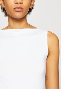 Hvid ærmeløs top, glat stof, bred halsudskæring, tætsiddende design. Har en minimalistisk stil uden mønstre eller synlige detaljer.