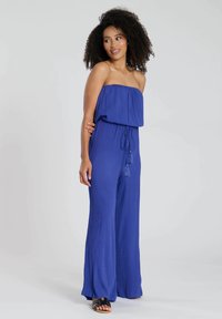 Mono azul sin tirantes con un cuerpo suelto, cintura atada y pantalones de pierna ancha. La tela tiene una textura arrugada y presenta borlas decorativas.