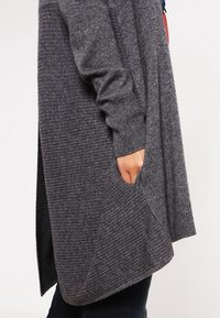 Cardigan gris en maille présentant une coupe longue, une texture côtelée, des poches latérales et un tombé subtil.
