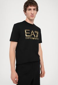 Black cotton t-shirt featuring a large gold EA7 Emporio Armani logo on the front. Round neckline and short sleeves. Smooth texture.